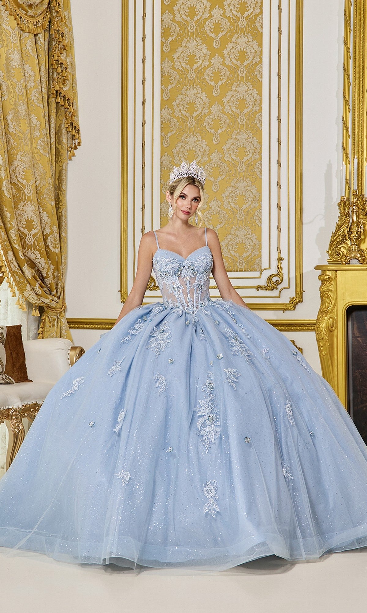 A woman in the Quinceanera Dress 5073 By Dancing Queen, featuring floral embroidery in dusty blue, and a tiara stands in an ornate gold and white room.